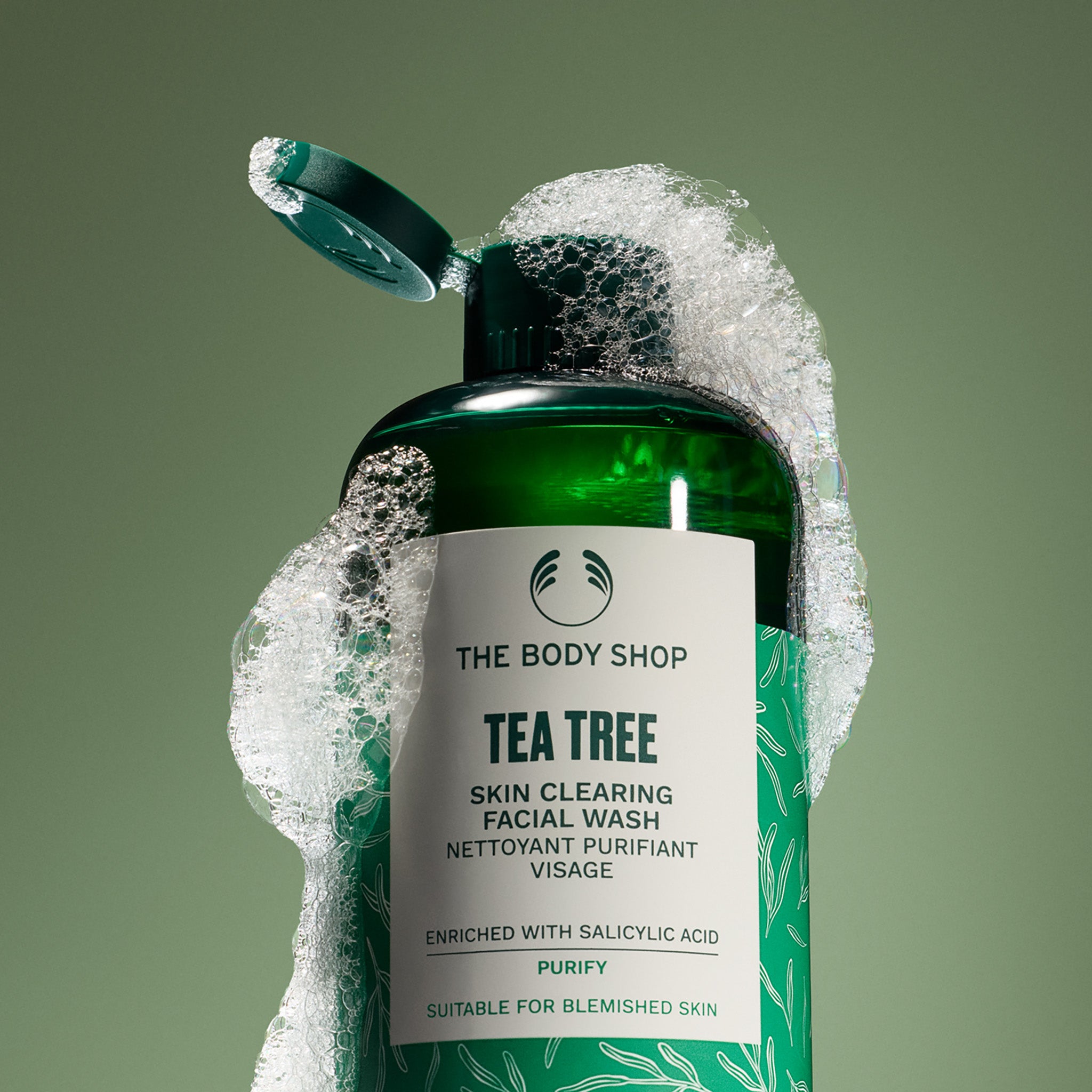 Green bottle of The Body Shop Tea Tree Skin Clearing Facial Wash with soap bubbles on a green background